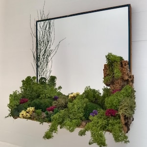 Op de afbeelding: Een rechthoekige spiegel met een donkerblauw frame, versierd met een naturalistische presentatie. De onderste rand heeft een plank van groen mos, kleine kleurrijke bloemen en een stuk schors. Een dunne, takachtige tak strekt zich omhoog uit.