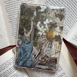 Può includere: Blocco acrilico rettangolare trasparente con un collage in stile vintage con una donna in abito blu, fiori e un disegno del sole. Il blocco poggia su libri aperti.