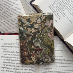 Puede incluir: Una funda transparente para teléfono con un diseño de bosque fantástico, que incluye una casa de hadas, setas y flores. El diseño incluye un borde de sello postal y la palabra "POSTAGE". Descansa sobre libros abiertos.