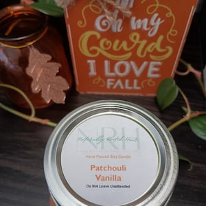May include: A silver-lidded jar candle with a label reading "Patchouli Vanilla." The candle sits on a wooden surface, with a decorative orange sign that says "Oh my Gourd I LOVE FALL." An orange glass jar and fall foliage are in the background.