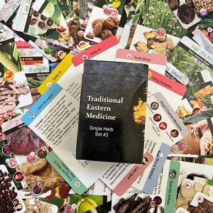 May include: A close-up of a black box with the text "Traditional Eastern Medicine Single Herb Set #3" on the front. The box is surrounded by a variety of colorful cards with images of herbs and text in a foreign language.