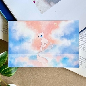 Può includere: Stampa artistica rettangolare con una creatura dei cartoni animati rosa chiaro e bianco con occhi blu, ambientata su un cielo pastello con una luna a mezzaluna. L'opera è esposta su un tavolo con libri aperti e vegetazione.