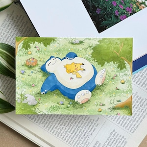 May include: A watercolour illustration shows a blue Snorlax and a yellow Pikachu resting on green grass. A small wooden crate and colourful eggs are scattered around the characters, under a tree. The artwork is on a white background.