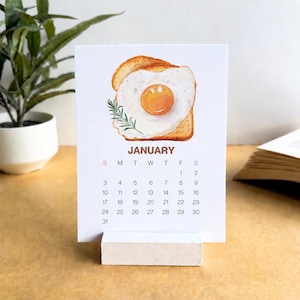 Puede incluir: Un calendario de escritorio blanco para enero que presenta una ilustración en acuarela de un huevo frito sobre una rebanada de pan tostado con una pequeña ramita de romero verde. La cuadrícula muestra los días del mes en negro sobre fondo blanco.