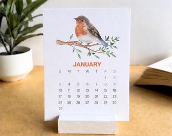 May include: A white paper desk calendar for the month of January, featuring a watercolour illustration of a robin perched on a leafy branch. The calendar displays the days of the week and dates in a grid format, supported by a rectangular white base.