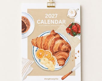Peut inclure: Un calendrier 2027 intitulé A YEAR OF BREAKFAST | WATERCOLOR PAINTINGS illustré avec un croissant, des tranches d'orange, des rondelles de banane, une tasse de café, un bol de fraises et un couteau à beurre sur fond beige avec une pince dorée.