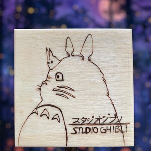 May include: A wooden music box with a hand-drawn image of Totoro from Studio Ghibli. The box is light brown and the image is in black and white.