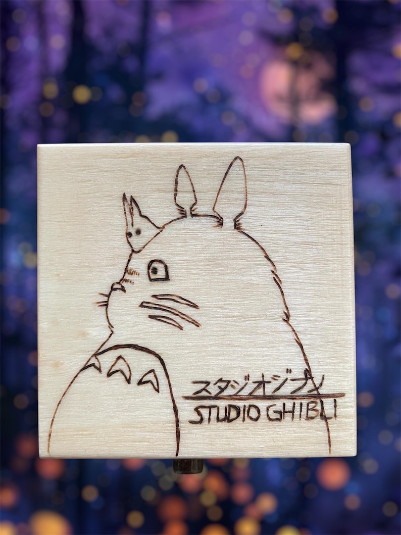 May include: A wooden music box with a hand-drawn image of Totoro from Studio Ghibli. The box is light brown and the image is in black and white.