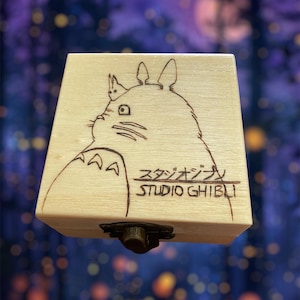 May include: A wooden music box with a wood-burned image of Totoro from Studio Ghibli. The music box is closed and has a small metal latch.