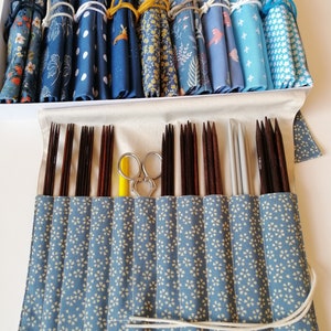 Könnte beinhalten: Eine blaue und weiße Stoffrolle mit einem weißen Kordelverschluss. Die Rolle enthält eine Reihe von Stricknadeln und eine gelbe und silberne Schere.