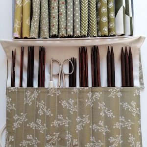 Könnte beinhalten: Ein Bastelset mit einer Vielzahl von Stricknadeln und Zubehör. Die Nadeln sind dunkelbraun und in einem Roll-Up-Etui mit floralem Muster angeordnet. Das Set enthält auch eine silberne Schere und Stoffbündel in Grün- und Weißtönen.