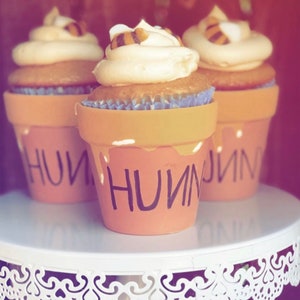 May include: Three cupcakes decorated with a bee design and topped with buttercream frosting. The cupcakes are displayed on a white tiered cake stand with a lace trim. The cupcakes are decorated with the word "HONEY" written on the side.