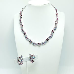 May include: A silver necklace and earrings set with a delicate design of pink and iridescent crystals. The necklace features a curved shape with a floral motif. The earrings are small and feature a similar floral design.