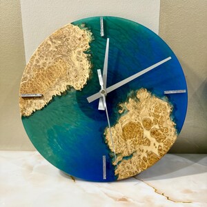 Puede incluir: Reloj de pared redondo con un dise&ntilde;o de resina epoxi azul y verde, con detalles de madera de ra&iacute;z de color marr&oacute;n claro. El reloj tiene manecillas plateadas y marcadores plateados en las posiciones de las 12, 3, 6 y 9 en punto. La esfera tiene unos 30 cm de di&aacute;metro.