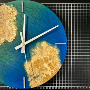 Puede incluir: Un reloj de pared redondo con un dise&ntilde;o &uacute;nico. La esfera del reloj presenta un fondo de resina azul y verde con un patr&oacute;n de vetas de madera. Las manecillas del reloj son blancas y los marcadores de hora son plateados. El reloj mide aproximadamente 25 cm de di&aacute;metro.