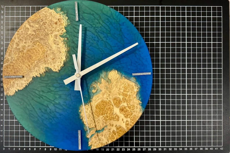 Puede incluir: Un reloj de pared redondo con un dise&ntilde;o &uacute;nico. La esfera del reloj presenta un fondo de resina azul y verde con un patr&oacute;n de vetas de madera. Las manecillas del reloj son blancas y los marcadores de hora son plateados. El reloj mide aproximadamente 25 cm de di&aacute;metro.