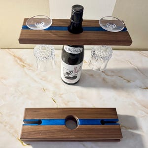 May include: A wooden wine caddy with a blue resin stripe, holding a wine bottle and two wine glasses. The caddy has a dark wood finish and a circular hole for the bottle. The wine bottle has a white label with Japanese text.