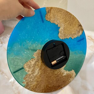 Puede incluir: Un reloj de pared redondo con un dise&ntilde;o de resina azul y marr&oacute;n. La esfera del reloj presenta un patr&oacute;n azul similar al oc&eacute;ano con acentos marrones similares a la madera. La parte posterior del reloj tiene un mecanismo de reloj negro con una bater&iacute;a Duracell.
