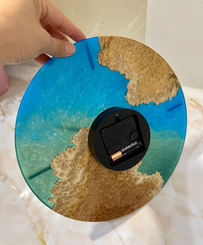 Puede incluir: Un reloj de pared redondo con un dise&ntilde;o de resina azul y marr&oacute;n. La esfera del reloj presenta un patr&oacute;n azul similar al oc&eacute;ano con acentos marrones similares a la madera. La parte posterior del reloj tiene un mecanismo de reloj negro con una bater&iacute;a Duracell.