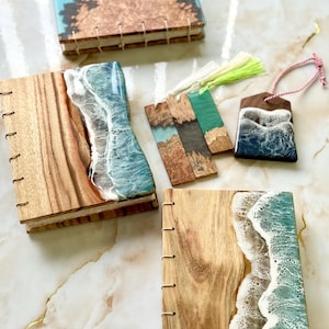 May include: A selection of handcrafted wooden notebooks and bookmarks. The notebooks display wood grain and resin designs with ocean wave and landscape themes. The bookmarks have colourful resin and wood accents, with a tassel and string.