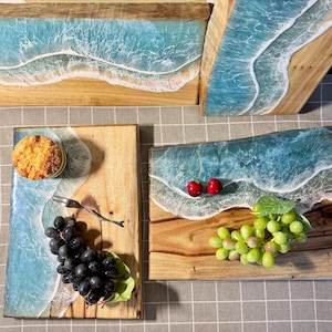 Op de afbeelding: Houten serveerplanken met een uniek oceaan golf ontwerp. De planken hebben een lichtbruine houten basis met een blauw-wit hars golfpatroon. Sommige planken zijn versierd met druiven, kersen en een klein gebakje.