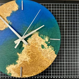 Puede incluir: Un reloj de pared redondo con un fondo de resina azul y verde, acentuado con un patr&oacute;n de vetas de madera marr&oacute;n claro. El reloj tiene manecillas plateadas y marcadores rectangulares plateados en las posiciones de las 12, 3, 6 y 9.