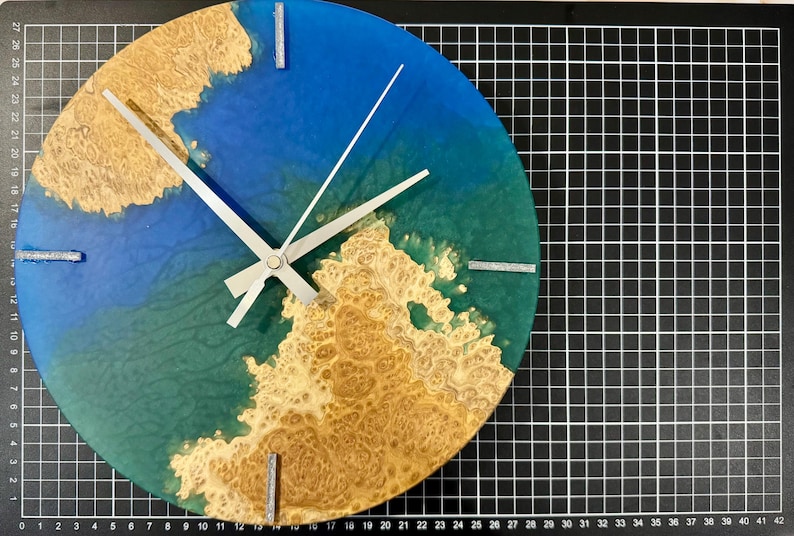 Puede incluir: Un reloj de pared redondo con un fondo de resina azul y verde, acentuado con un patr&oacute;n de vetas de madera marr&oacute;n claro. El reloj tiene manecillas plateadas y marcadores rectangulares plateados en las posiciones de las 12, 3, 6 y 9.