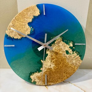 Puede incluir: Reloj de pared redondo con fondo de resina azul y verde, acentuado con vetas de madera marr&oacute;n claro. Agujas y marcadores plateados. Una pieza decorativa &uacute;nica, de unos 30 cm de di&aacute;metro.