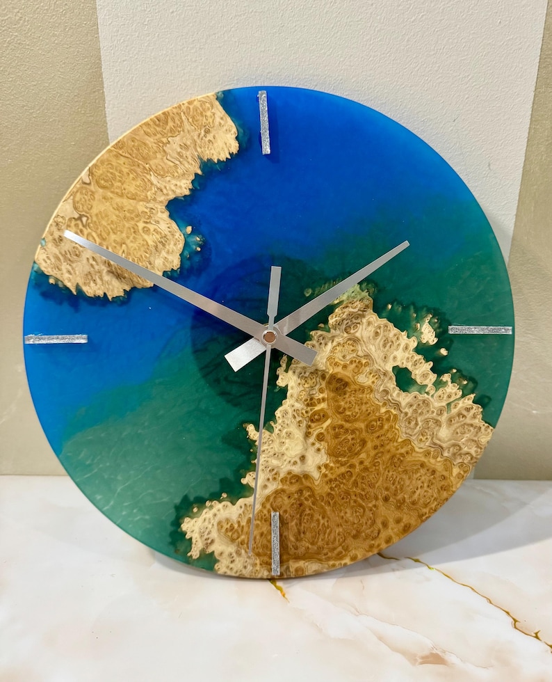 Puede incluir: Reloj de pared redondo con fondo de resina azul y verde, acentuado con vetas de madera marr&oacute;n claro. Agujas y marcadores plateados. Una pieza decorativa &uacute;nica, de unos 30 cm de di&aacute;metro.