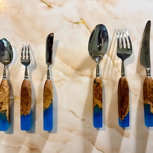 May include: A set of six stainless steel utensils: two forks, two spoons, and two knives. The handles are made of wood and blue resin. The wood has a natural grain pattern, and the resin is a vibrant blue color.