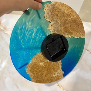 Puede incluir: Un reloj redondo con un dise&ntilde;o de resina azul y marr&oacute;n. La esfera del reloj presenta un patr&oacute;n azul arremolinado que se asemeja al agua, con detalles de vetas de madera de color marr&oacute;n. El reloj tiene un mecanismo de reloj negro en la parte posterior.