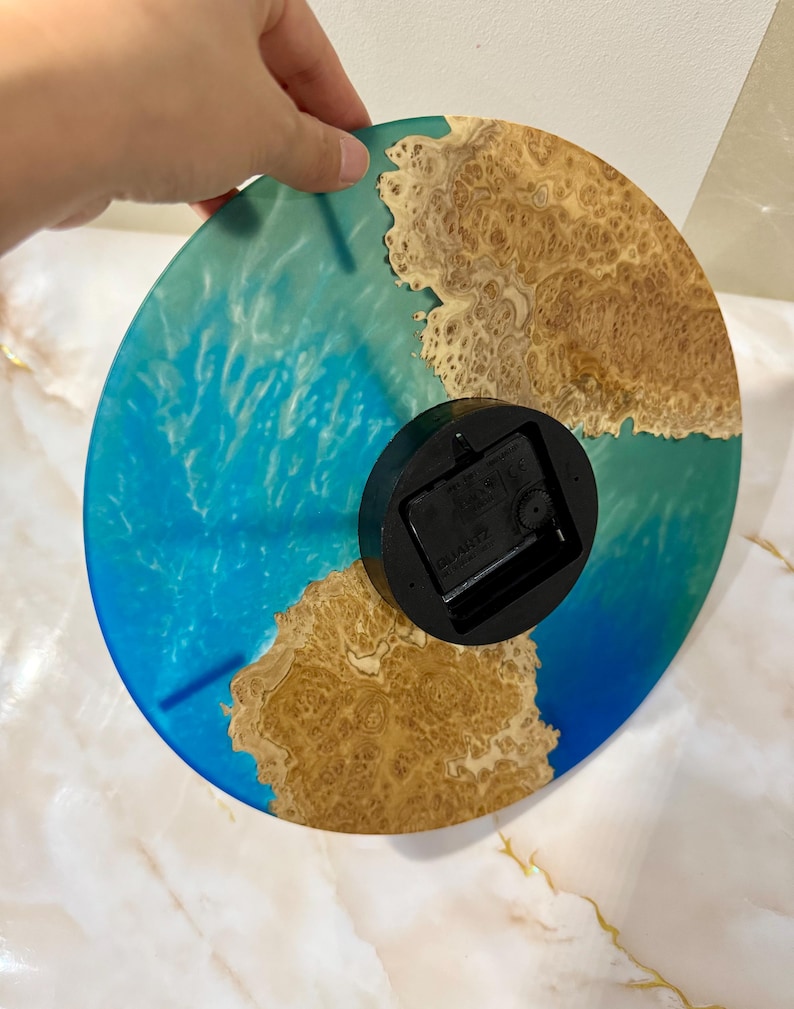 Puede incluir: Un reloj redondo con un dise&ntilde;o de resina azul y marr&oacute;n. La esfera del reloj presenta un patr&oacute;n azul arremolinado que se asemeja al agua, con detalles de vetas de madera de color marr&oacute;n. El reloj tiene un mecanismo de reloj negro en la parte posterior.