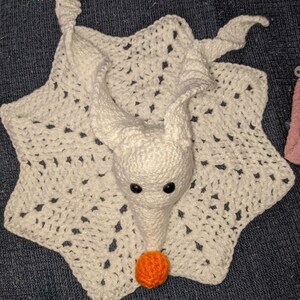 May include: White crocheted baby blanket with a crocheted ghost design. The ghost has black eyes and an orange nose.