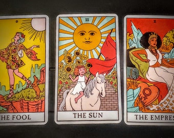 Peut inclure: Trois cartes de tarot, Le Fou, Le Soleil et L'Impératrice. La carte du Fou montre une personne vêtue d'une tenue colorée marchant vers une falaise avec un chien. La carte du Soleil montre un enfant chevauchant un cheval blanc avec un drapeau rouge et un soleil avec un visage. La carte de l'Impératrice montre une femme assise sur un trône avec une couronne et un cœur avec un symbole féminin.