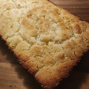 Op de afbeelding: Een rechthoekig brood met een goudbruine, gebarsten en getextureerde korst. Het bovenoppervlak vertoont een rustiek, onregelmatig uiterlijk met diepe scheuren en een lichtbruine kleur, wat wijst op een goed gebakken, stevig brood.