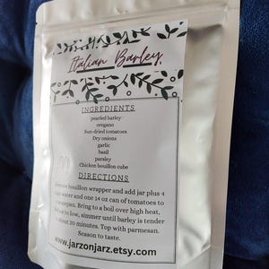 May include: A white stand-up pouch containing Italian Barley with a label listing ingredients including pearled barley, oregano, sun-dried tomatoes, dry onions, garlic, basil, parsley, and a chicken bouillon cube, with cooking directions included.