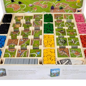 May include: A board game organiser box containing various game components including square cardboard tiles with landscape illustrations, wooden meeples in black, blue, green, yellow, pink, and red, and a small white cloth bag, all arranged in wooden dividers.