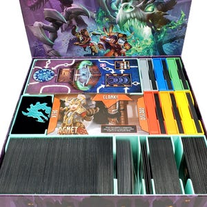 May include: Open board game box with various components. Includes cards, game boards, and dividers in a purple box. The box art features fantasy characters and a dragon. Text on the cards reads "Deck", "Clank!", and "Discard".