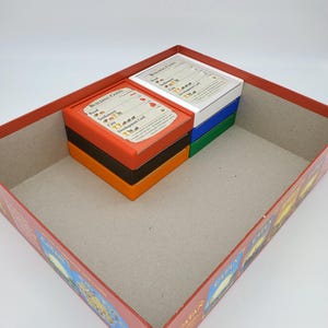 May include: The inside of a Catan game box with the game pieces organized in their designated compartments. The compartments are labeled with the game's building costs, including the cost of roads, settlements, cities, and development cards.