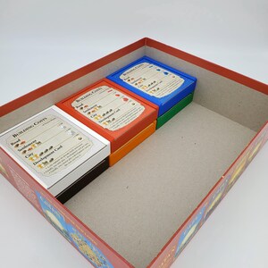 May include: A red game box with a cardboard insert for storing game components. The insert has four compartments, each containing a stack of cards with different colored backs. The cards are labeled "Building Costs" and show the cost of different game pieces in terms of resources.
