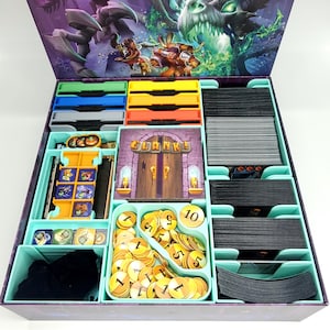 May include: A board game organizer with various compartments and game components. The box contains stacks of cards, game tokens, and colorful trays. The lid features fantasy artwork with a dragon and characters. The game title "Clank!" is visible.