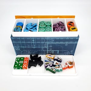 Könnte beinhalten: Ein Brettspiel-Set mit verschiedenen bunten Spielsteinen, darunter Würfel, Spielmarken und Spielfiguren. Die Spielsteine werden in einem geteilten weißen Tablett und einer blauen Box mit einem Bauplan-Design aufbewahrt.
