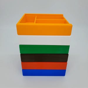 May include: A stack of six colorful plastic storage boxes. The boxes are orange, white, green, brown, red, and blue. The boxes are all the same size and shape, and they are stacked on top of each other.
