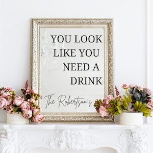 May include: A gold framed mirror with the text "You look like you need a drink" and the text "The Robertson's" below it. The mirror is sitting on a white mantelpiece with two pink flower arrangements on either side.