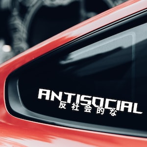May include: White "ANTISOCIAL" sticker with Japanese characters, applied to the window of a red car. The word "ANTISOCIAL" is in a bold, sans-serif font, with the Japanese translation beneath.