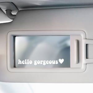 Könnte beinhalten: Ein weißer Sonnenblende für das Auto mit einem Spiegel und einem weißen Vinyl-Aufkleber mit der Aufschrift "hello gorgeous" mit einem Herzen.