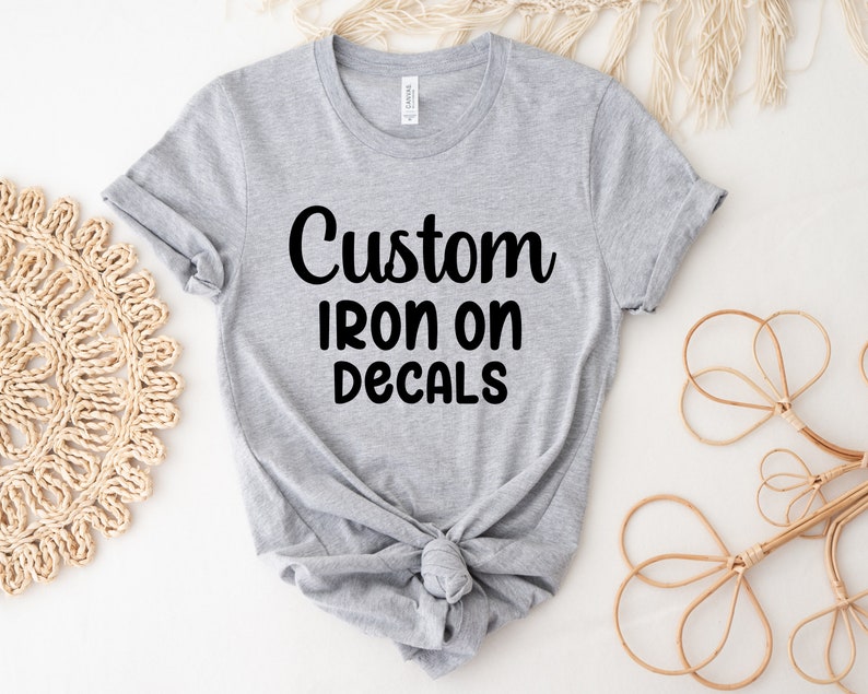 May include: A gray t-shirt with the words "Custom Iron On Decals" in black script. The shirt is knotted at the waist. Decorative woven and floral elements are in the background.