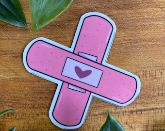 May include: A pink bandage sticker in a cross shape with a white border. The sticker has a small heart in the center. The sticker is on a wooden surface with green leaves.
