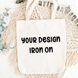 May include: Cream-colored tote bag with the text "YOUR DESIGN IRON ON" in black. The bag has two handles and is made of a sturdy fabric. The background includes decorative elements like macrame and greenery.
