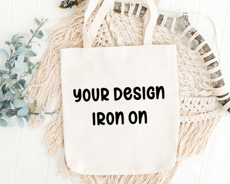May include: Cream-colored tote bag with the text "YOUR DESIGN IRON ON" in black. The bag has two handles and is made of a sturdy fabric. The background includes decorative elements like macrame and greenery.